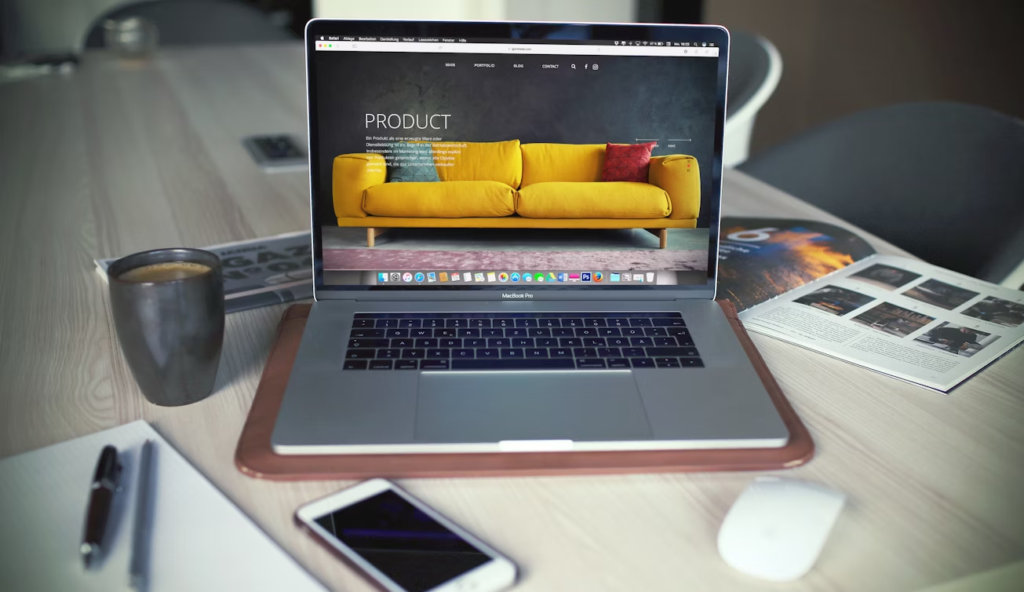 MacBook Pro displaying an e-commerce website with a yellow sofa on a modern workspace desk, featuring a smartphone, coffee cup, mouse, notepad, and magazine—ideal setup for online business and digital marketing.
